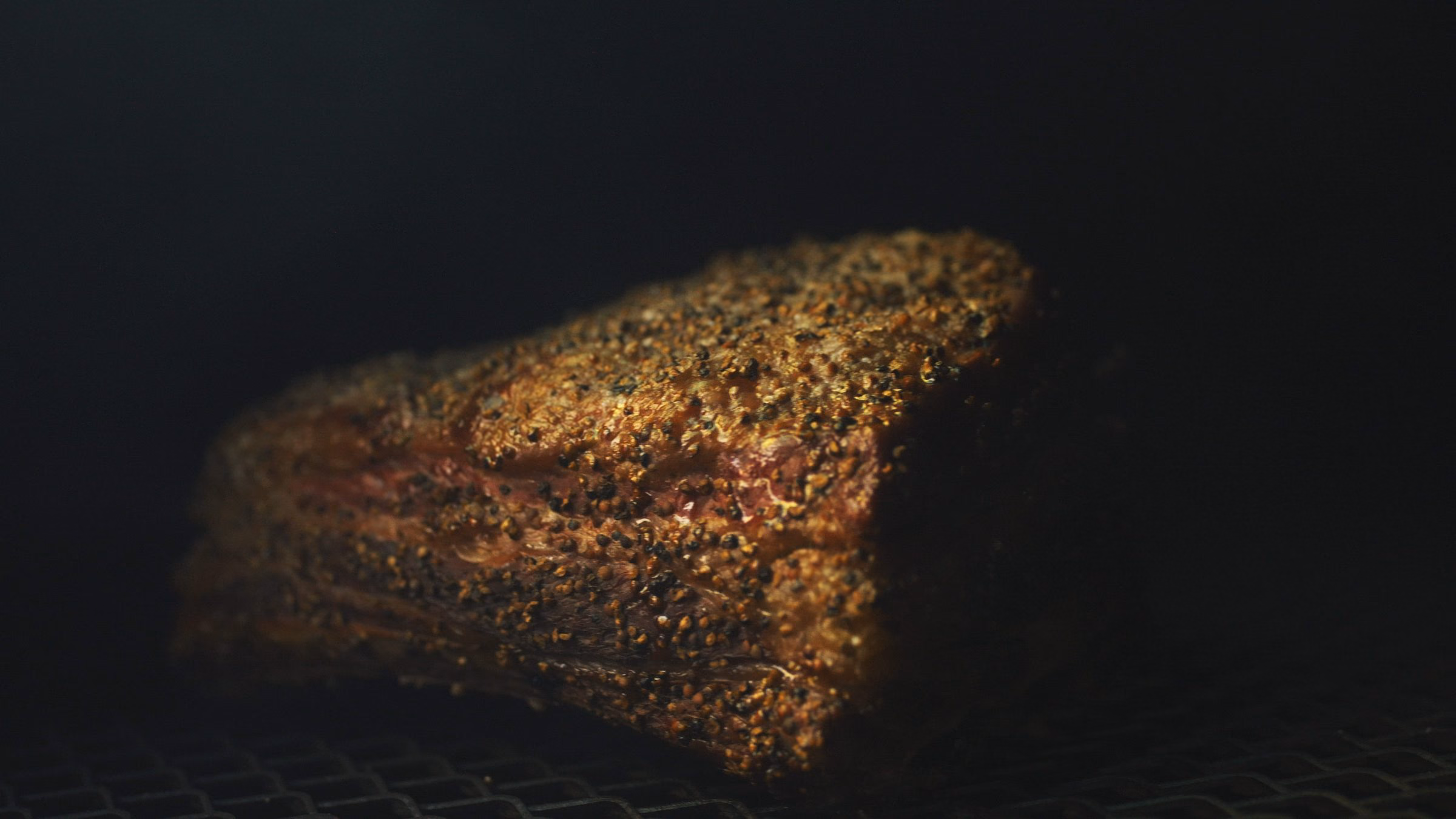 Pièce de bœuf couverte d’un rub poivré sur la grille du smoker, fond sombre