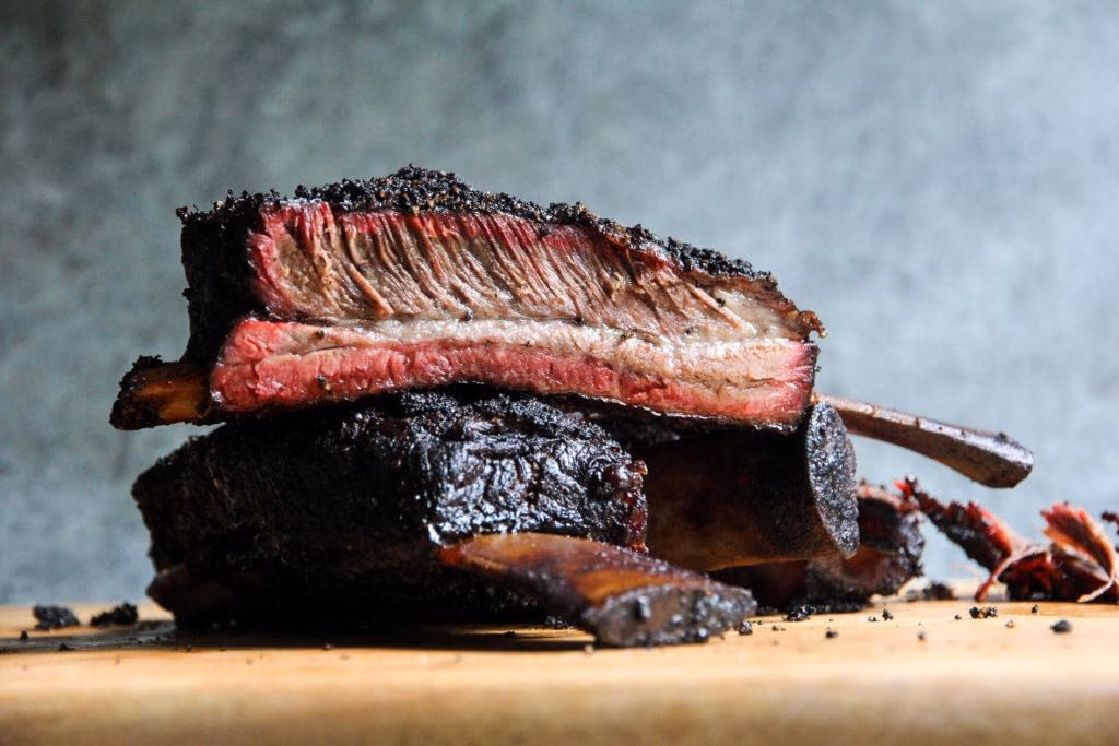 Ribs de bœuf fumés empilés sur une planche, bark noir poivré et anneau de fumée rosé en coupe